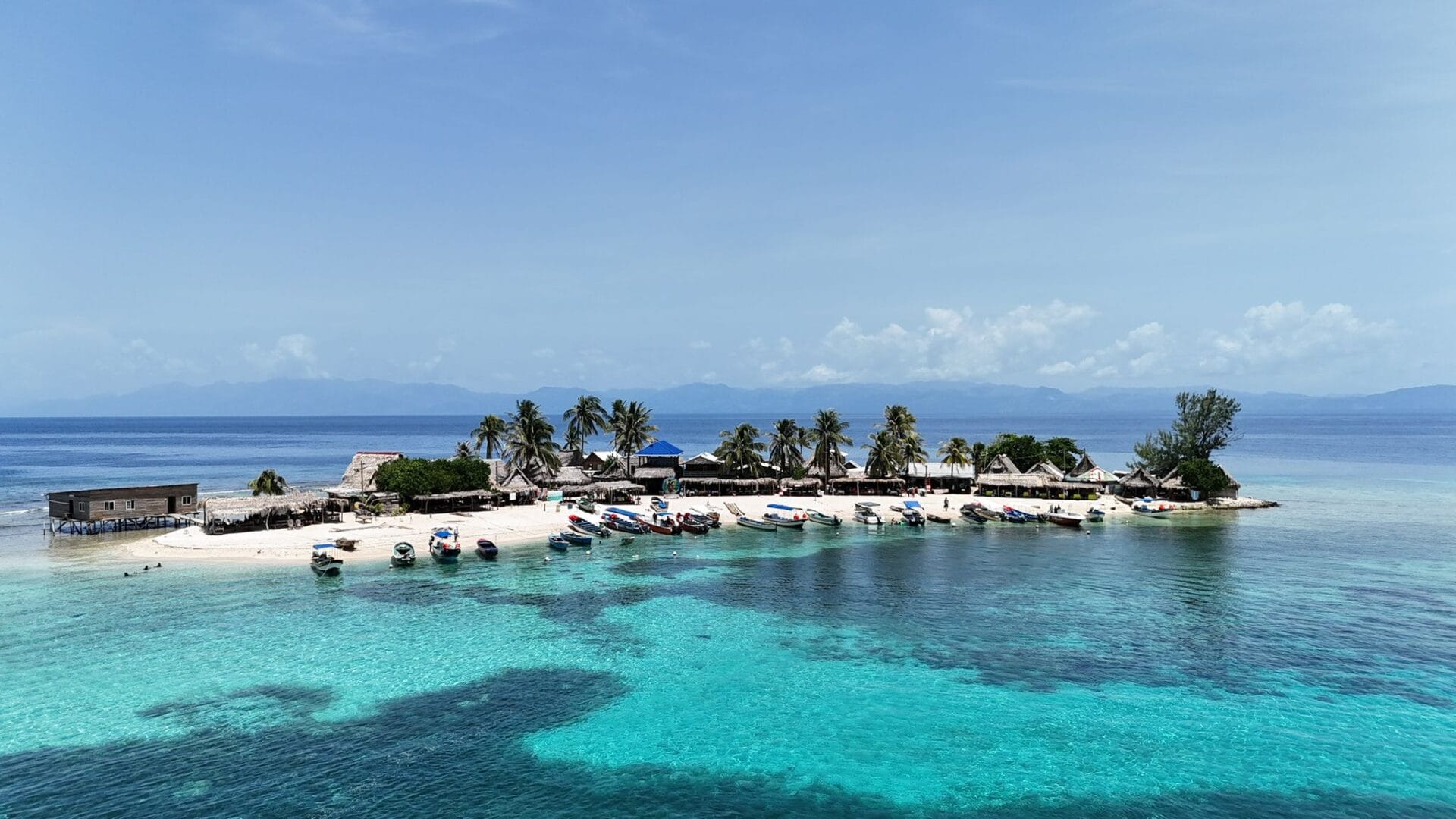 A scenic view of a small tropical island in Roatan, Honduras, with turquoise waters, numerous boats docked on the white sandy beach, palm trees, and thatched-roof structures against a backdrop of distant mountains and a clear blue sky.