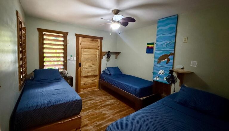 A cozy bedroom with three twin beds featuring blue bedding and wooden frames, situated in a rustic room with wooden flooring and natural finished wood doors, located in Roatan, Honduras.