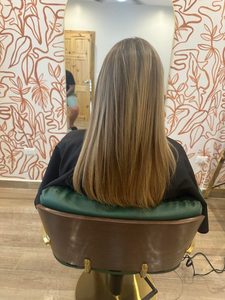 The back of a woman with long, straight blonde hair seated in a stylish salon chair facing a wall with patterned wallpaper on Roatan, Honduras. A mirror reflects a wooden door and a person in casual clothing sitting nearby.