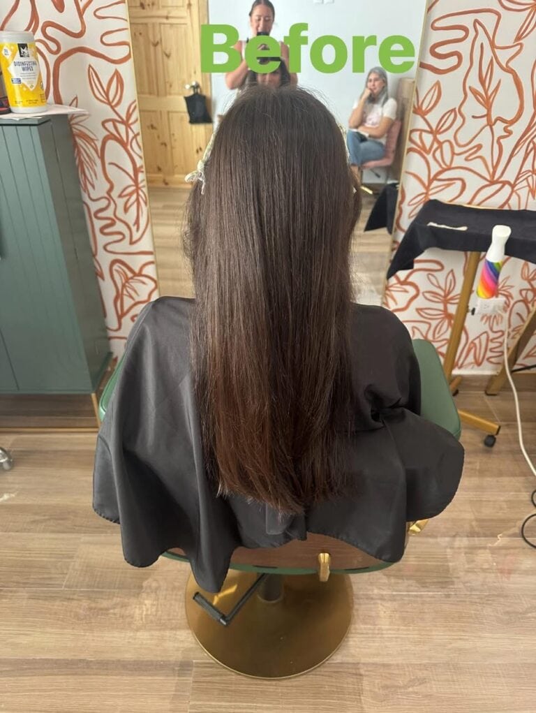 A woman with long brown hair seated in a hair salon on Roatan, Bay Islands, Honduras, before a haircut or hair treatment, with a large mirror reflecting her and the salon interior.