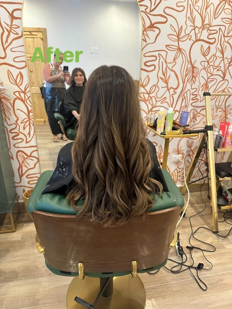Woman with long wavy brown hair sitting in a salon chair in Roatan, Honduras, after a hair treatment, with a stylist and client visible in the mirror reflection.