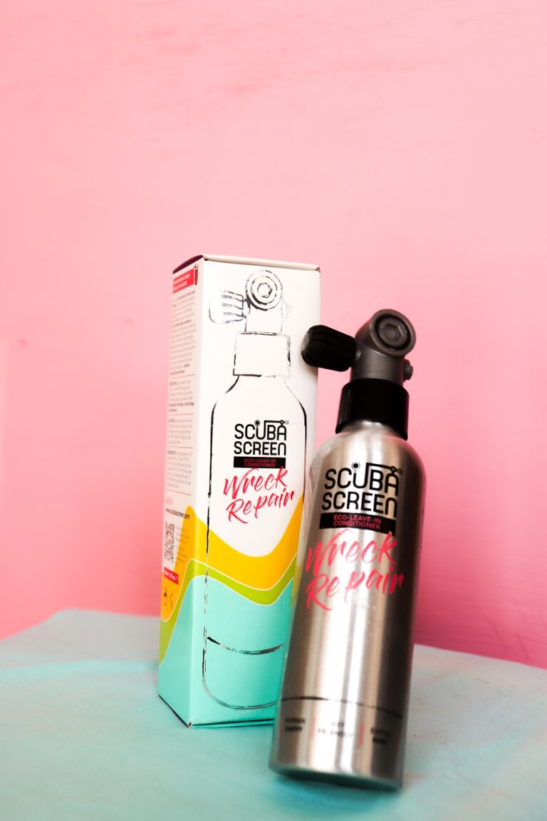A silver spray bottle of Wreck Repair eco-leave-in conditioner next to its colorful packaging against a pink background on Roatan in Honduras.