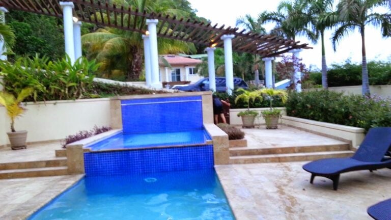 Tiered blue-tiled swimming pool with a raised spa, pergola, palms, and lounge chairs at a resort in Roatan, Honduras.