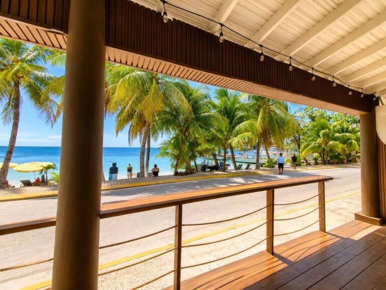 A covered wooden veranda overlooks palm trees, a beachfront walkway, and the Caribbean Sea in Roatan, Honduras.