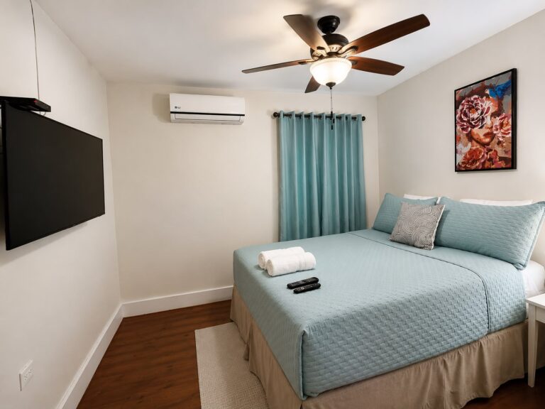 Bright Roatan, Honduras bedroom with a neatly made bed, teal curtains, ceiling fan, air conditioner, wall-mounted television, and tropical artwork.