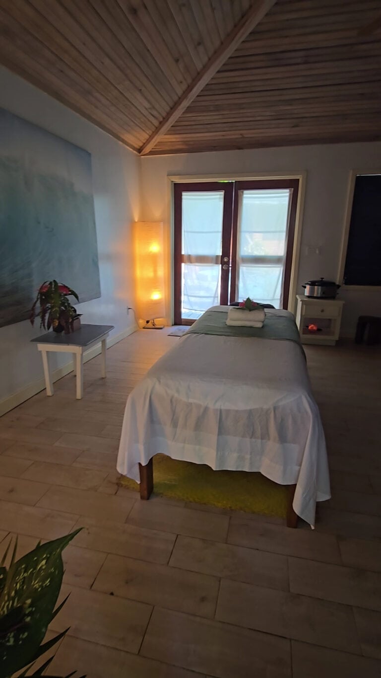 Tranquil massage room with a treatment table, wood-paneled ceiling, tropical plants, and ocean-facing doors in Roatan, Honduras.