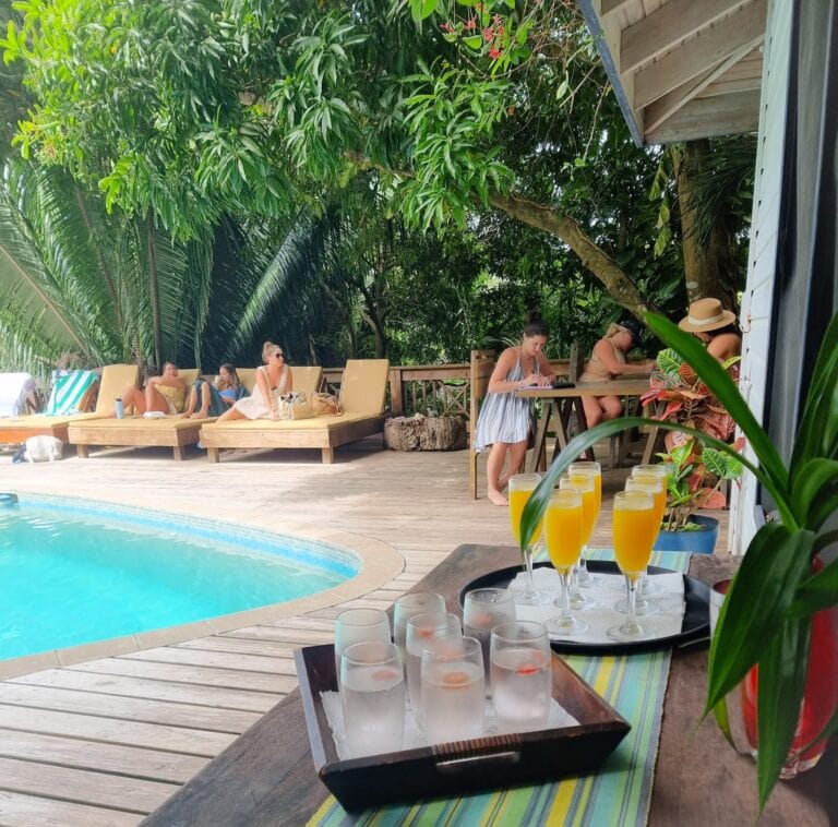Guests relax beside a turquoise swimming pool with tropical greenery and drinks on a wooden deck in Roatan, Honduras.