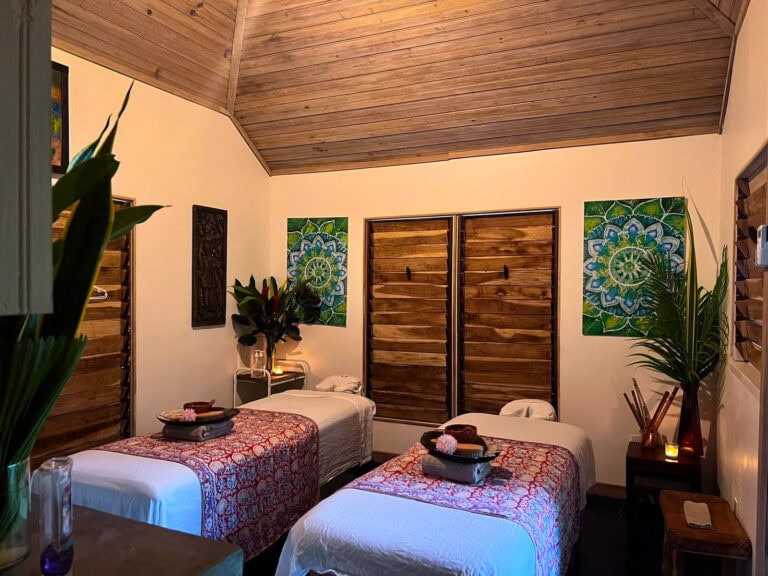 Twin bedroom in Roatan, Honduras, featuring two beds with patterned runners, tropical plants, colorful wall art, and wooden double doors.
