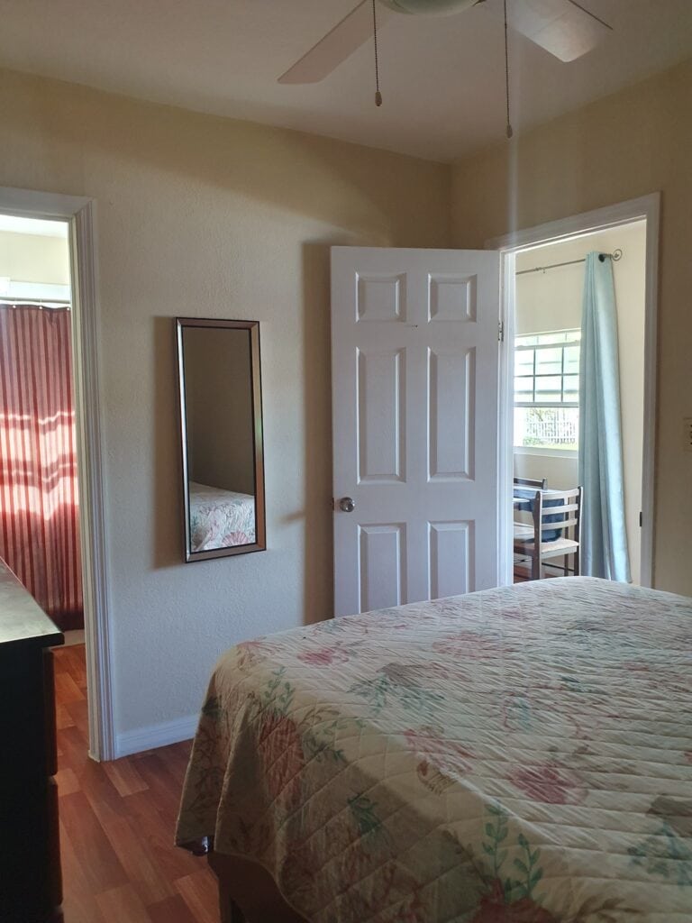 A cozy bedroom in a Roatan, Honduras vacation rental with a bed, mirror, and nearby window with a blue curtain.