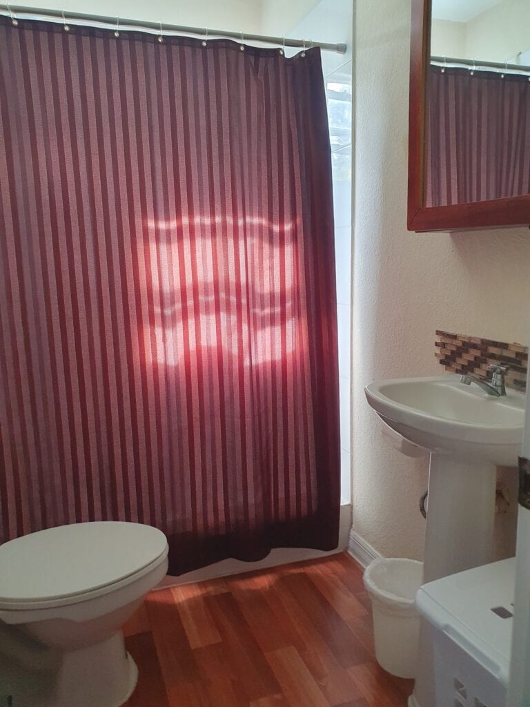 Bathroom on Roatan with a striped shower curtain, a white toilet, a small wall-mounted sink, and a mirror with a wooden frame reflecting part of the room.