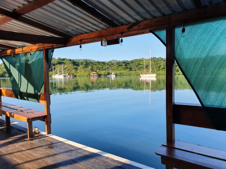 A wooden and metal covered dock on Roatan in Honduras overlooks calm water with sailboats anchored nearby and lush green hills in the background.