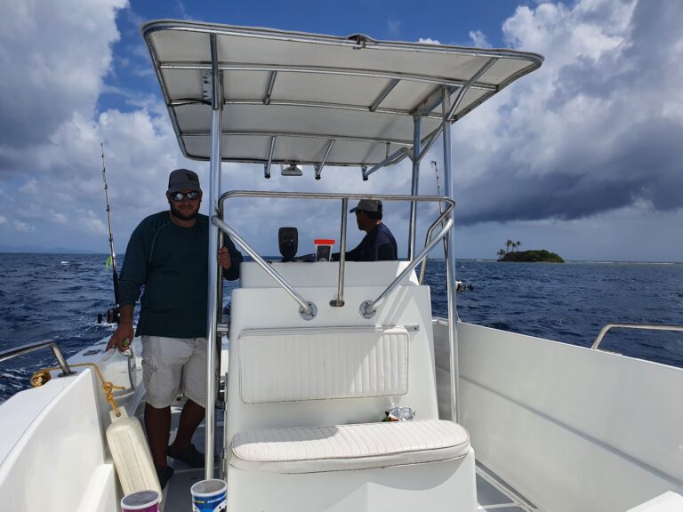 Two men are on a boat in the waters near Roatan, Honduras, with an island featuring palm trees visible in the background under a partly cloudy sky.