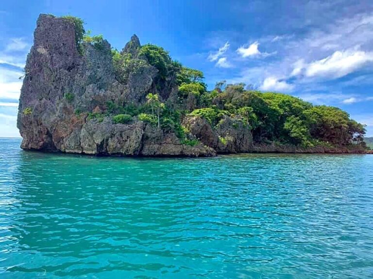 A rocky island covered with lush green vegetation rises from the turquoise waters off the coast of Roatan in Honduras, under a partly cloudy blue sky.