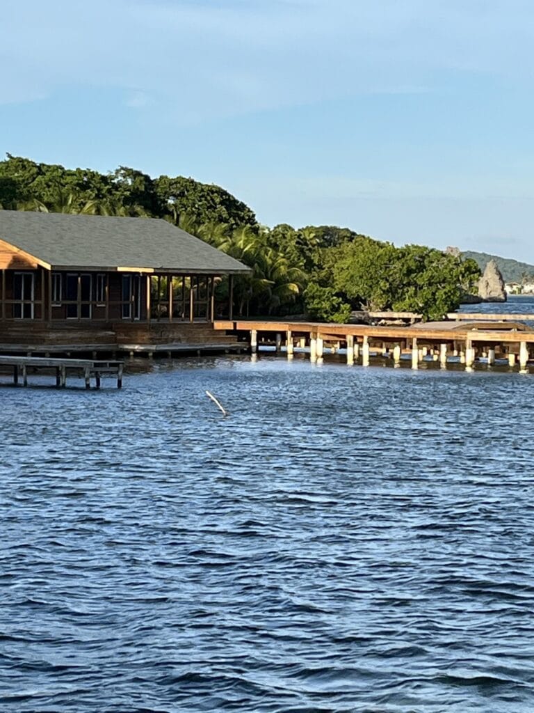 Overwater wooden bungalows with thatched roofs are built on stilts above the clear blue waters of Roatan in the Bay Islands, Honduras, surrounded by lush green tropical trees and distant rocky formations.