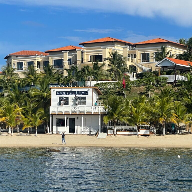 Luxury beachfront residential complex with multiple cream-colored buildings featuring red-tiled roofs and large windows on Roatan, Honduras, surrounded by lush palm trees under a clear blue sky.