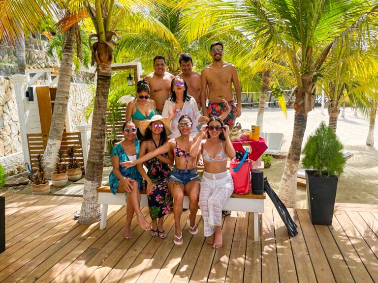A group of ten friends enjoying a tropical beach party under palm trees on Roatan, Honduras with sandy shores and lush greenery in the background.