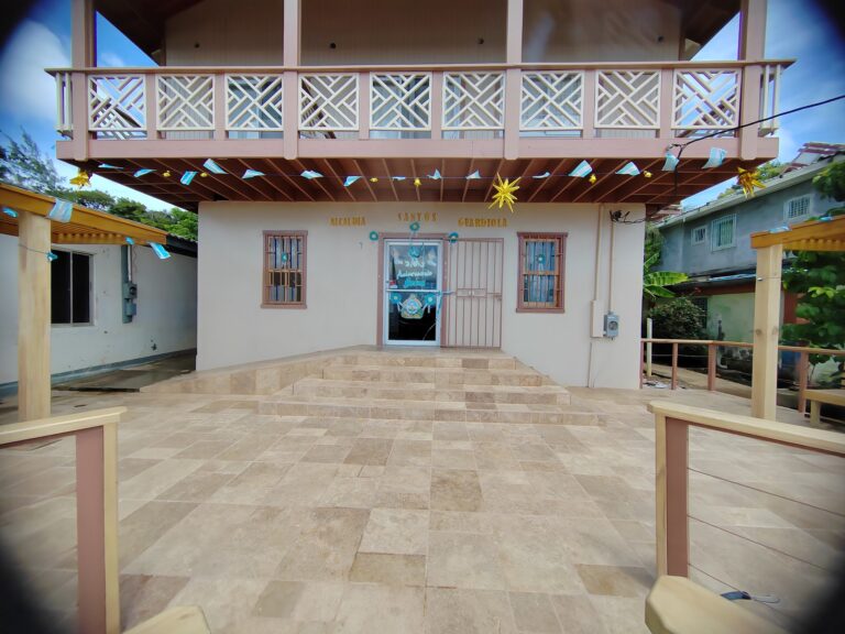 A two-story building in Roatan, Honduras, features a decorative balcony on the upper level with a patterned railing, festive hanging decorations below, and a sign reading "Academia Santos Guaridola" above the entrance.