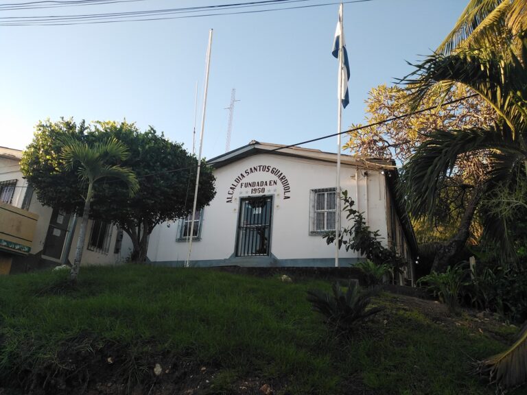 A white building with black window bars and a sign indicating it is the Alcadia Santos Guardiola, founded in 1950, is surrounded by lush green trees and tropical plants on Roatan, Honduras.