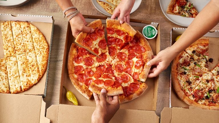 Multiple people are sharing and serving different types of pizza from pizza boxes on a wooden table, with the setting indicating a casual dining experience in Roatan, Honduras.