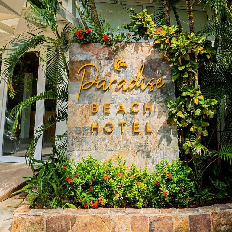 A decorative stone wall with the words "Paradise Beach Hotel" illuminated in yellow cursive letters, surrounded by lush tropical plants and flowers on Roatan, Honduras.