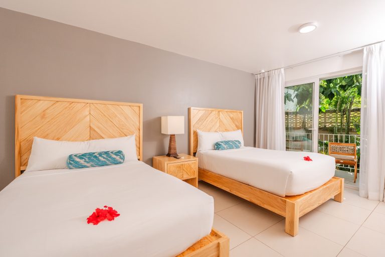 Two neatly made twin beds with wooden headboards and pillows are placed in a bright room with large windows overlooking lush greenery in Roatan, Honduras.