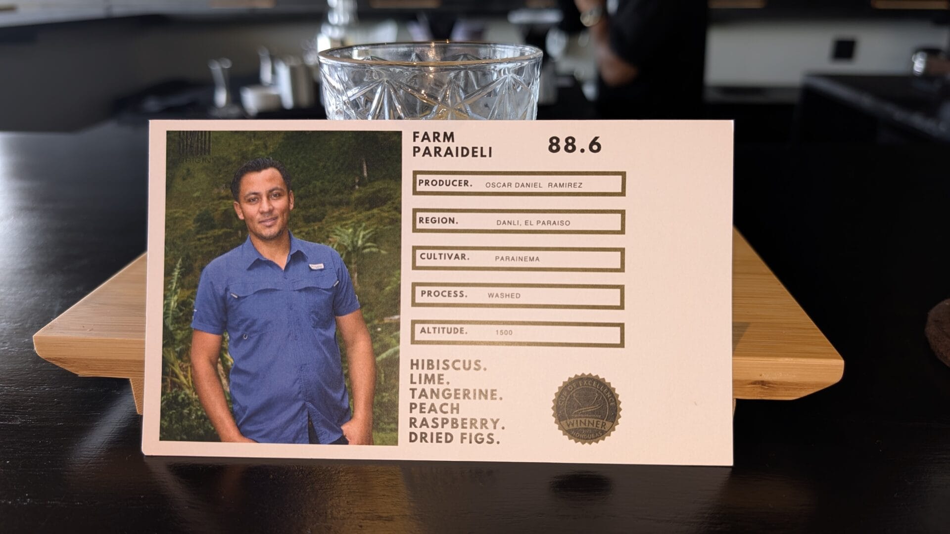 A close-up of a coffee cupping sheet featuring a photo of a man in a blue shirt with a lush green background, placed on a wooden table in Roatan, Honduras.