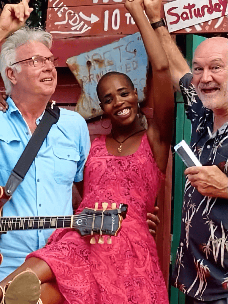 Three smiling musicians pose with a guitar outside a colorful venue in Roatan, Honduras.