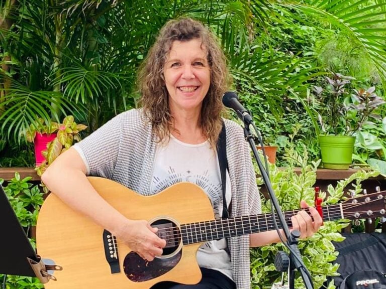 A woman with curly hair playing an acoustic guitar and singing into a microphone outdoors amidst lush green tropical plants on Roatan in Honduras.