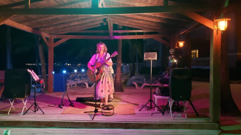 A woman performs live music with a guitar on a wooden stage under a thatched roof at night in Roatan, Honduras, with pink and orange lighting, surrounded by lounge chairs and tropical decor.