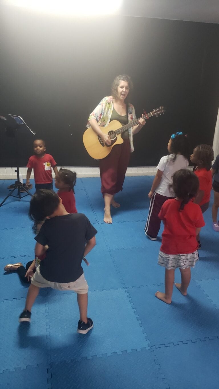 A woman playing guitar entertains children during a music class or performance on Roatan, Honduras, with kids gathered around on blue interlocking foam mats in an indoor setting.