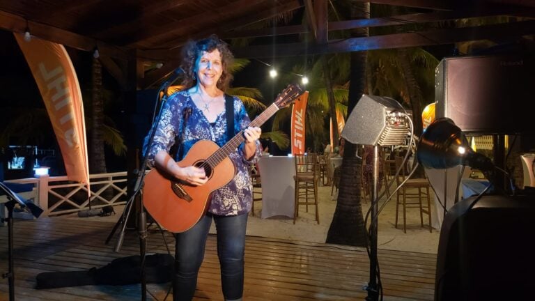 A woman playing acoustic guitar and singing on an outdoor stage at night in Roatan, Honduras, with palm trees, wooden deck, and decorative lighting in the background.