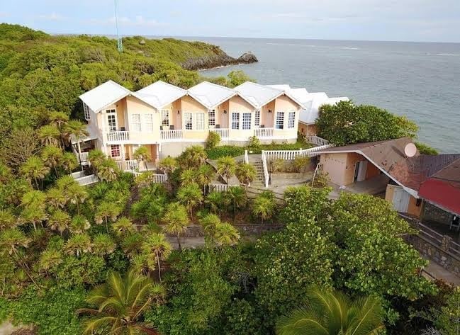 A white multi-unit beachfront resort building with large windows and balconies is surrounded by lush green tropical vegetation on Roatan in the Bay Islands of Honduras, with the ocean visible in the background.