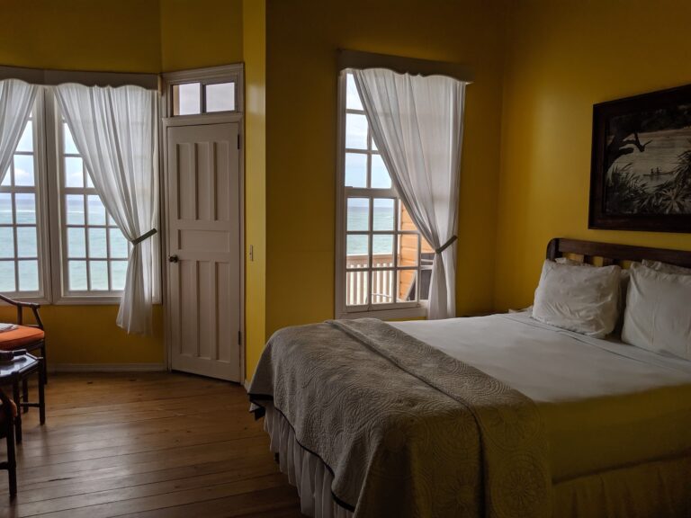 A bright yellow bedroom in Roatan, Honduras, features two large windows with white curtains revealing ocean views, with a bed partially visible on the right side.