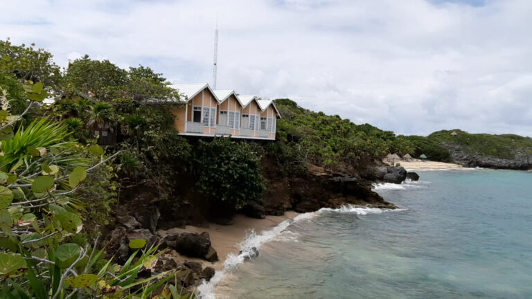 A row of wooden beach cottages with steep roofs are built on a lush green hillside overlooking the ocean on Roatan, Honduras, with a sandy beach, rocks, and waves in the foreground and a partly cloudy sky above.