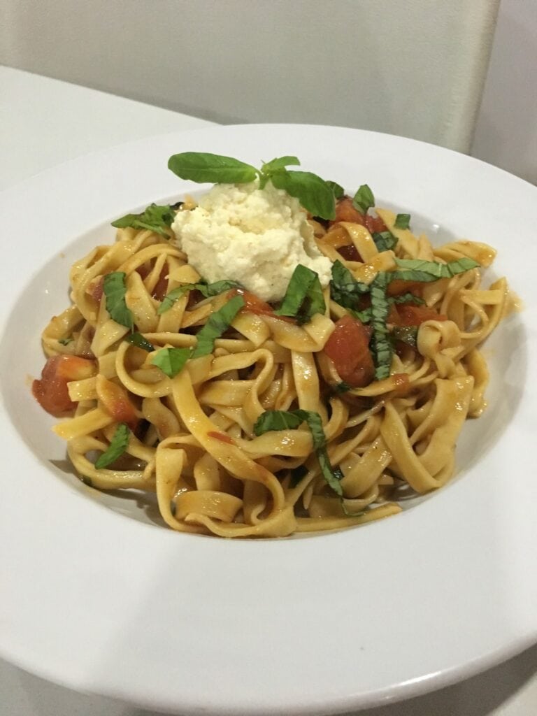A plate of pasta with cherry tomatoes, fresh basil, and ricotta cheese, presented on a white plate against a neutral background in Roatan, Honduras.