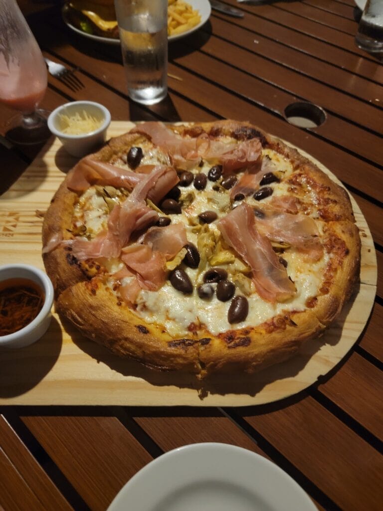 A fully baked pizza topped with ham, black olives, and cheese, served on a wooden platter with small bowls of dipping sauces, located at a restaurant in Roatan, Honduras.