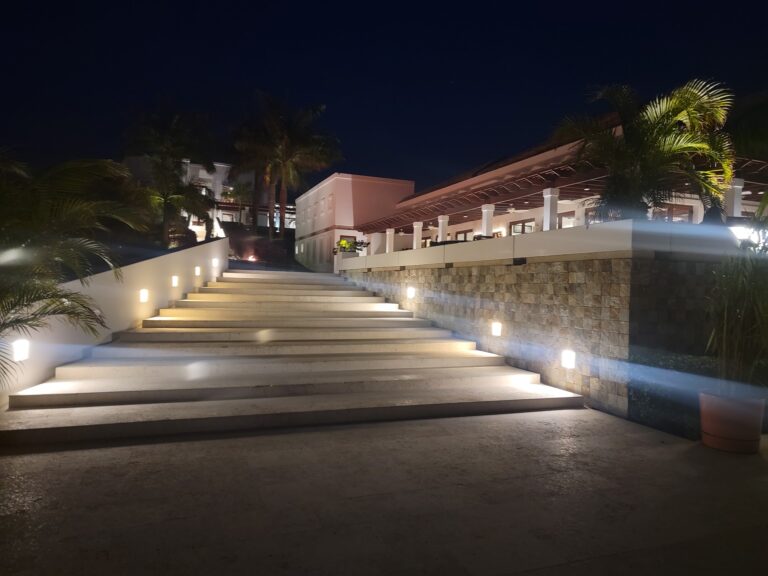 A modern, illuminated staircase leading up to a building with white walls and a wooden overhang, surrounded by palm trees on Roatan, Honduras at night.