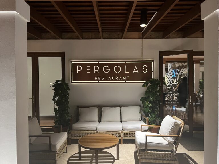 A seating area inside a restaurant on Roatan, Honduras, features white cushioned chairs, wooden tables, and greenery, with a sign displaying "Pergolas Restaurant" on the wall.