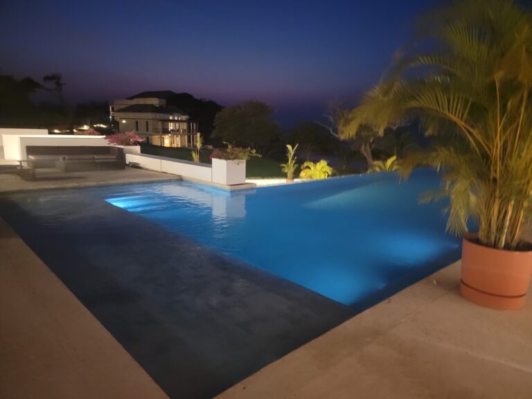 An illuminated swimming pool at night with palm trees and modern houses overlooking Roatan, Honduras, in the background.