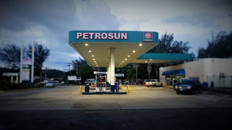 A Petrosun gas station with an illuminated canopy displaying the Petrosun logo is situated in Roatan, Honduras, surrounded by parked vehicles, with a cloudy sky overhead.