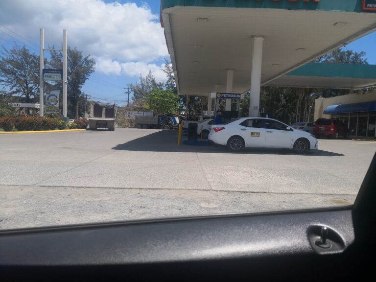 A Petrosun gas station in Roatan, Honduras, with white and red cars parked under a canopy, surrounded by trees and a cloudy sky.