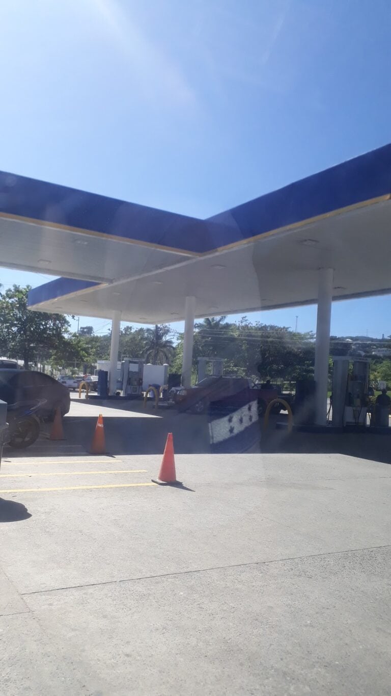 A gas station in Roatan, Honduras, with multiple fueling pumps, orange traffic cones, and cars parked nearby under a large canopy with a clear blue sky overhead.