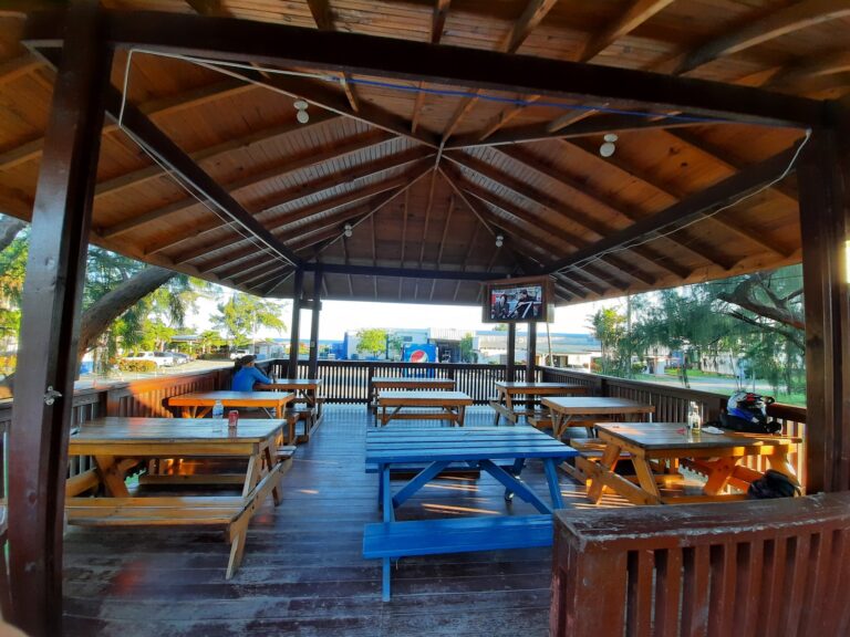 An open-air wooden pavilion in Roatan, Honduras, features picnic tables, string lights, and a small television, with trees and a parking lot visible in the background.