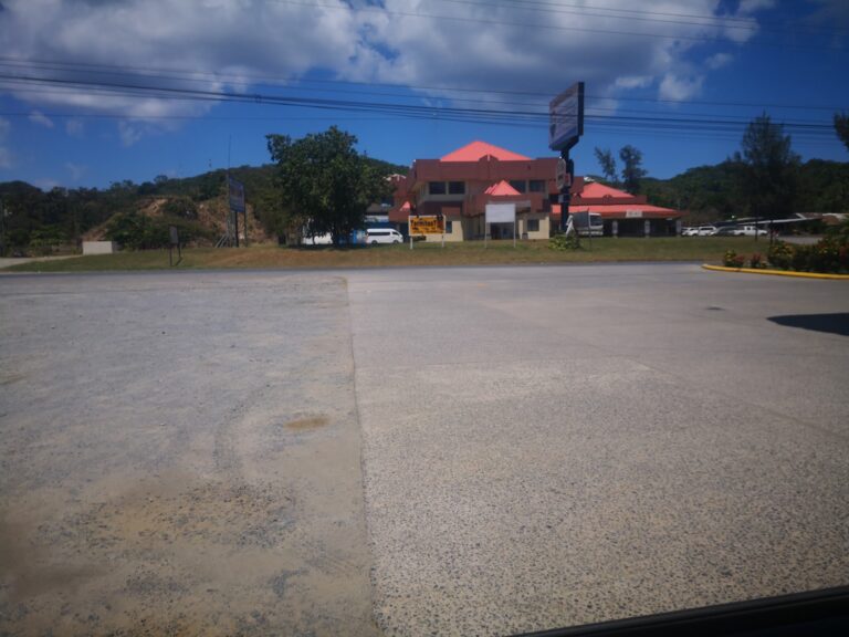 A building with a red roof and a beige exterior sits along a road in Roatan, Honduras, with parked white vehicles and a backdrop of green hills and cloudy blue sky.