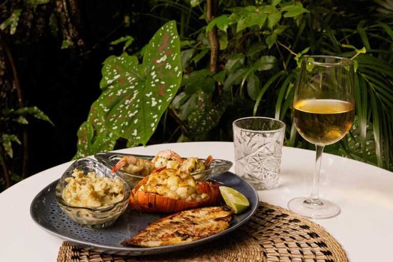 A seafood dish including lobster tail, shrimp, and fish served with mashed potatoes, a lime wedge, and a glass of white wine on a table surrounded by lush green tropical plants in Roatan, Honduras.