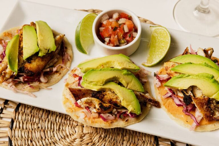 Three fish tacos topped with sliced avocado, served on a white platter with lime wedges, pico de gallo, and a glass of white wine, representing authentic cuisine from Roatan, Honduras.