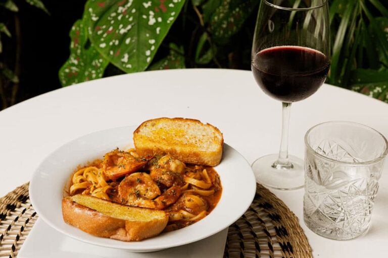 A plate of pasta with shrimp and sauce, served with two slices of garlic bread, a glass of red wine, and a glass of water on a white table with green plants in the background in Roatan, Honduras.