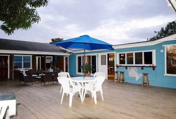 A wooden outdoor patio in Roatan, Honduras, features a white round table with four white chairs under a large blue umbrella, surrounded by a blue building with windows and doorways, and an area with black seating and bar stools.