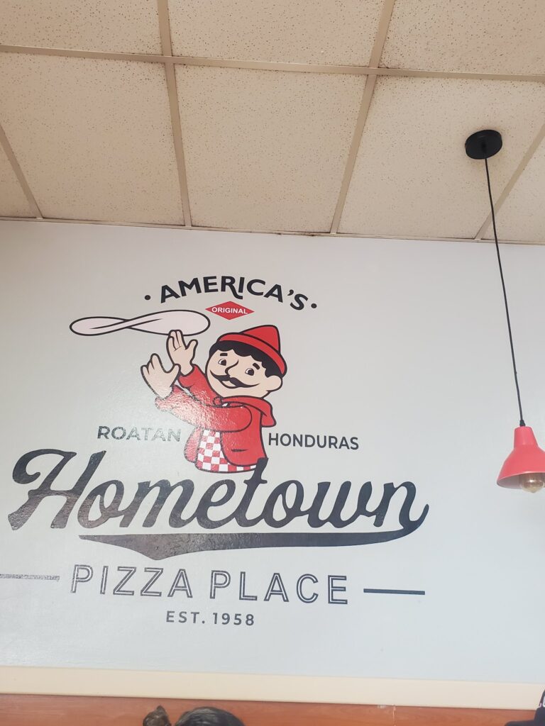 A sign displaying the name "Hometown Pizza Place" with a cartoon character wearing a red hat and checkered apron, and text indicating it is located in Roatan, Honduras, established in 1958.