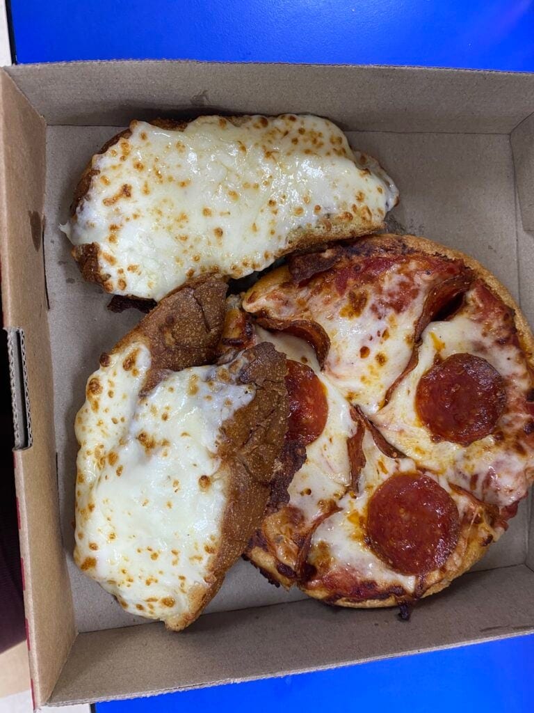 A cardboard box contains three slices of pizza with melted cheese, pepperoni, and a breaded fried mozzarella cheesestick, set against a blue background, representing food options available in Roatan, Honduras.
